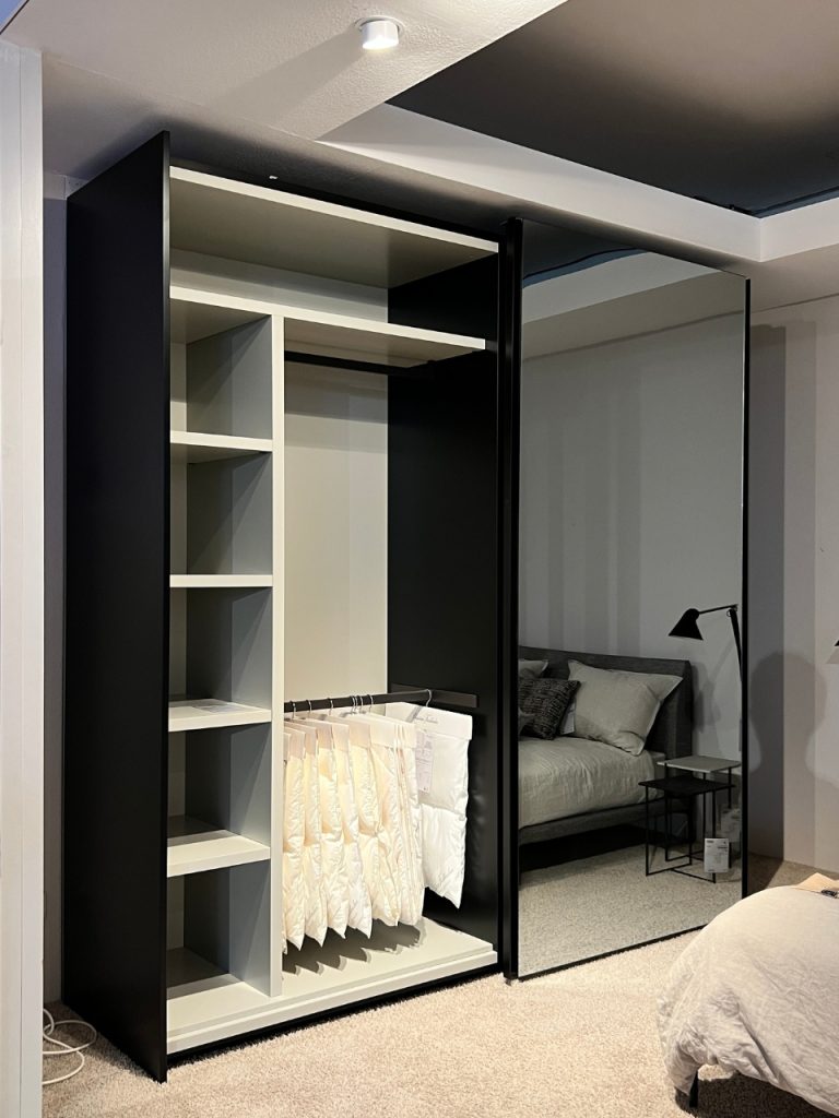 Open wardrobe with shelves and a hanging rack for white pants; a mirrored door reflects a gray bedroom with a bed and black floor lamp.