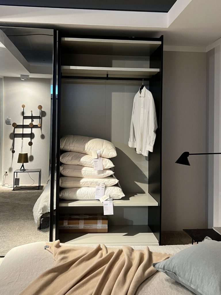 Open wardrobe showing a white shirt on a hanger and a stack of four pillows on the shelf with a bed visible in the foreground.