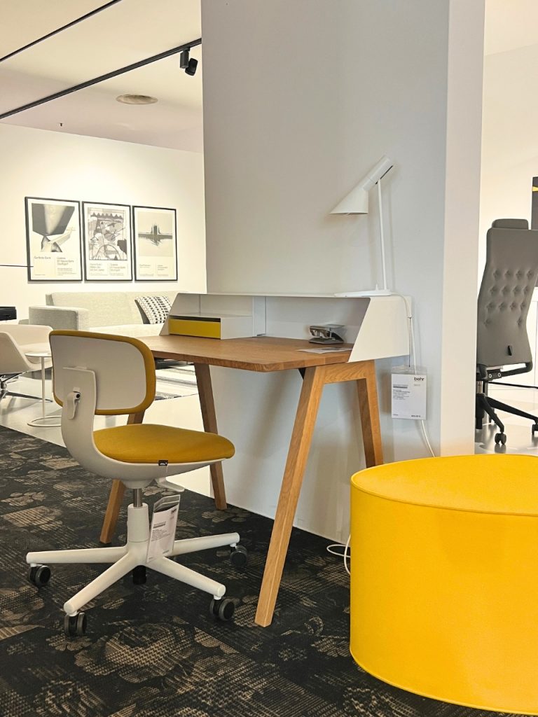 Modern showroom workspace: wooden desk with white storage on a white partition, yellow-cushioned chair, white chair base, and a bright yellow ottoman nearby.