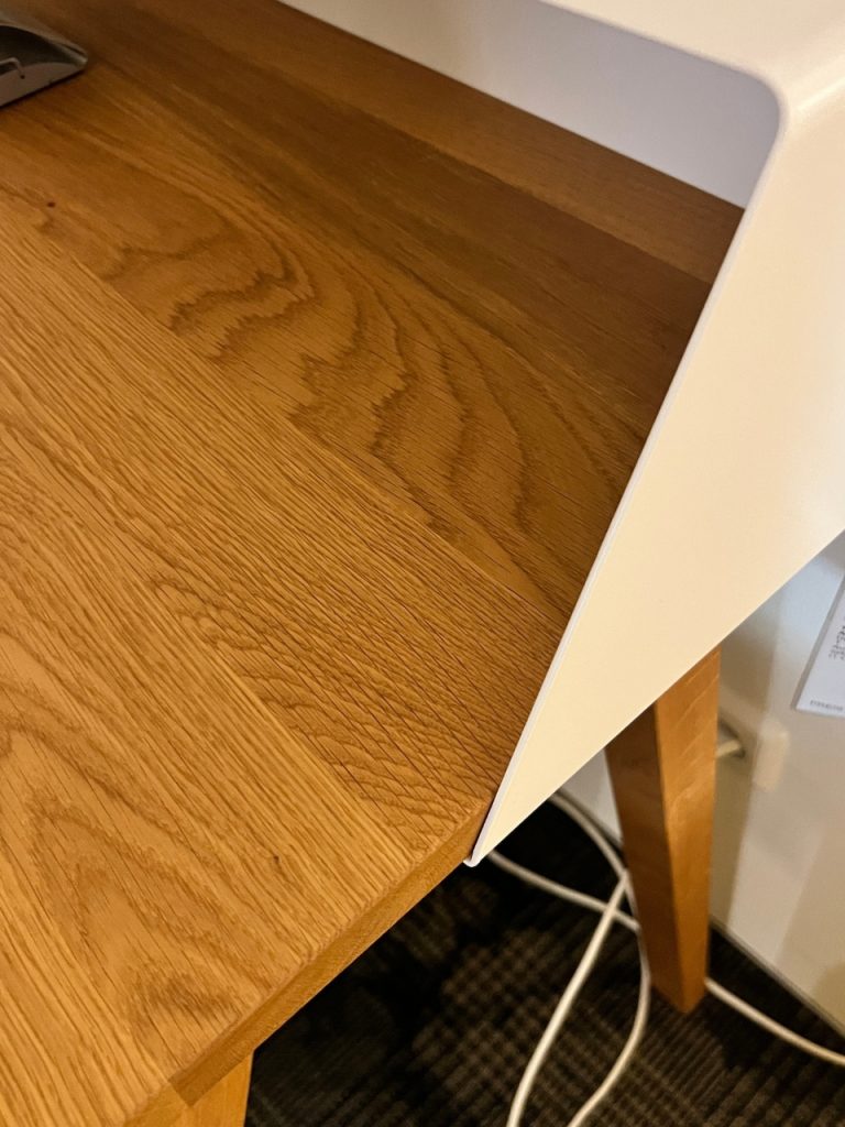 Corner of a wooden desk with a white edge panel, with cables on the carpet beneath.
