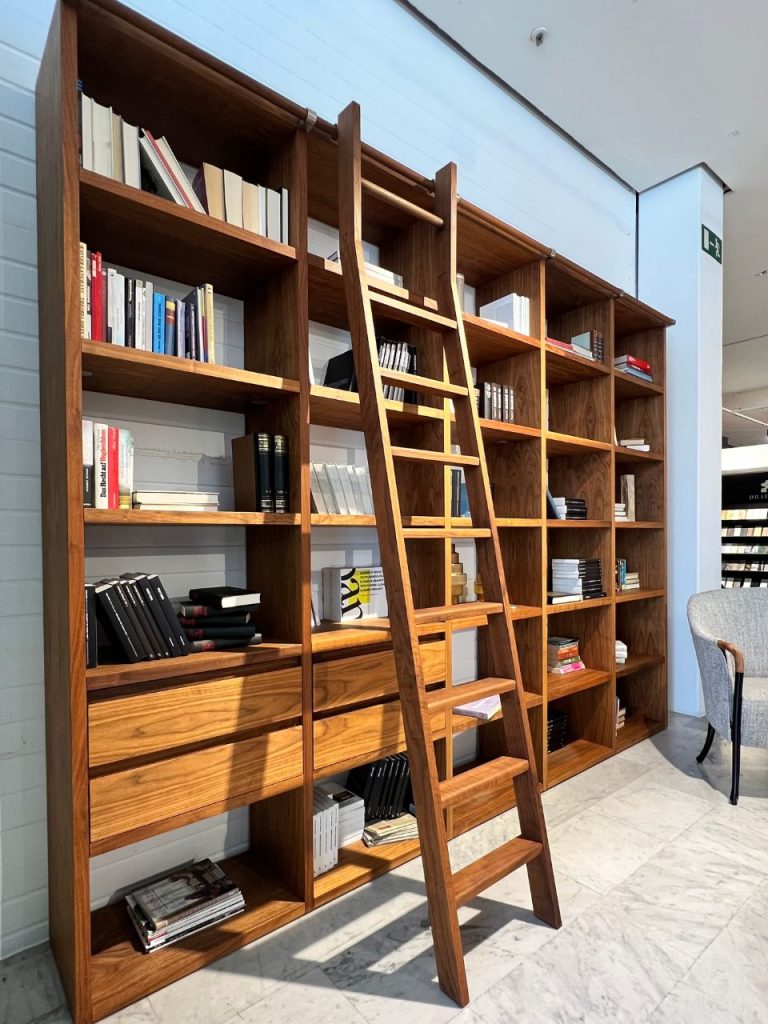 Tall wooden bookcases with a ladder spanning their shelves, filled with books in a bright, modern reading area or bookstore.