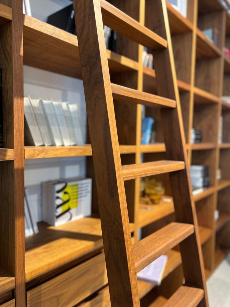 Wooden library shelves with a built-in ladder leaning against them, filled with books and magazines in a warm, modern setting