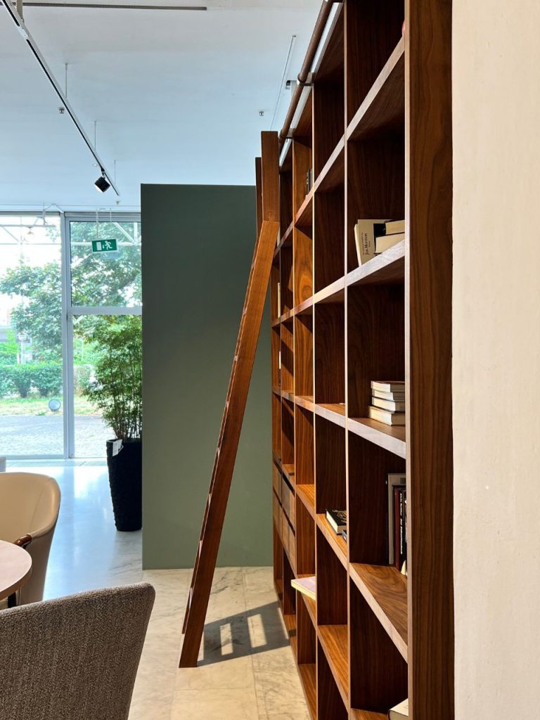 Tall wooden bookshelf with a leaning ladder in a bright reading area; glass doors, exit sign, and potted plant visible in the background.