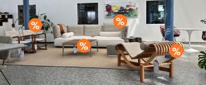 Furniture showroom with a sofa, chairs, and coffee table featuring orange discount badges on several pieces of furniture in a styled room setting.