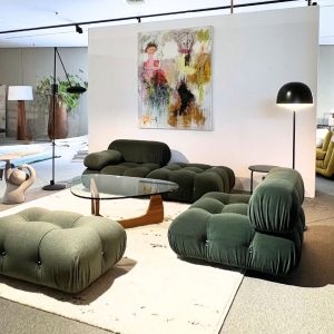 Green tufted modular sofas surround a round glass coffee table in a modern showroom, with an abstract painting on a white divider.