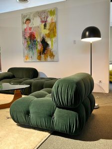 Green, plush, modular sofa with rounded sections in a contemporary showroom setting, paired with a glass coffee table and a black arc floor lamp beside an abstract art painting on the wall.