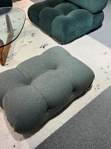 Gray tufted ottoman in the foreground with a dark green tufted ottoman behind it on a light rug beside a glass round coffee table.