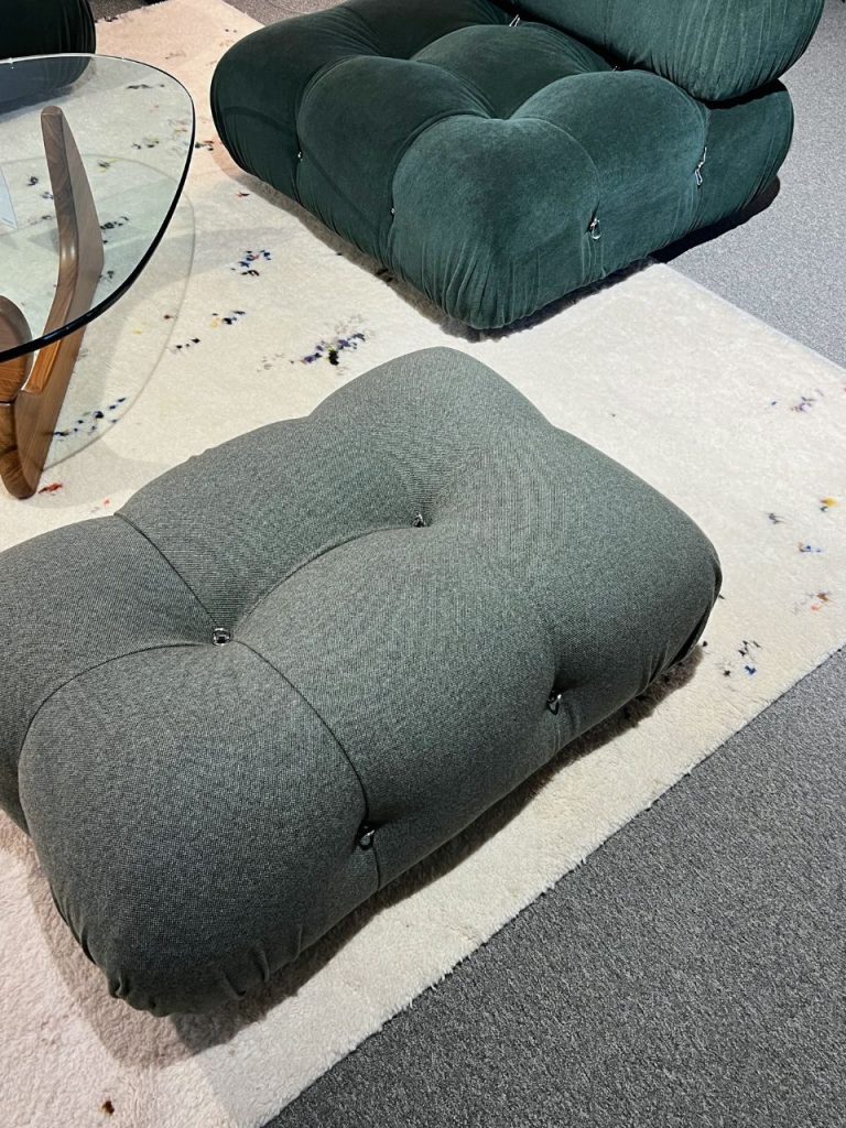 Gray tufted ottoman in the foreground with a dark green tufted ottoman behind it on a light rug beside a glass round coffee table.
