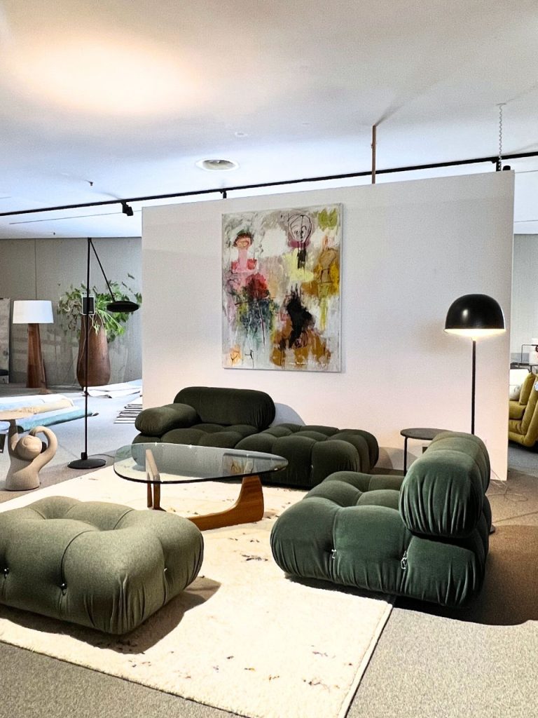 A modern lounge area with dark green tufted modular Sofas arranged around a glass coffee table in front of a white partition displaying an abstract painting.
