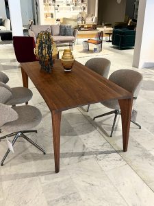 Wooden rectangular dining table with four grey upholstered chairs in a furniture showroom; decorative vases on the table and blurred living-room displays in the background.