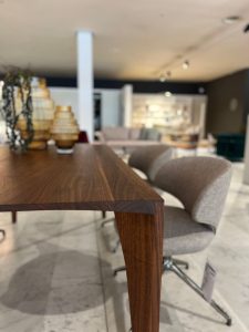 Close-up of a solid wood table edge in a modern showroom, with blurred chairs and display shelves in the background.