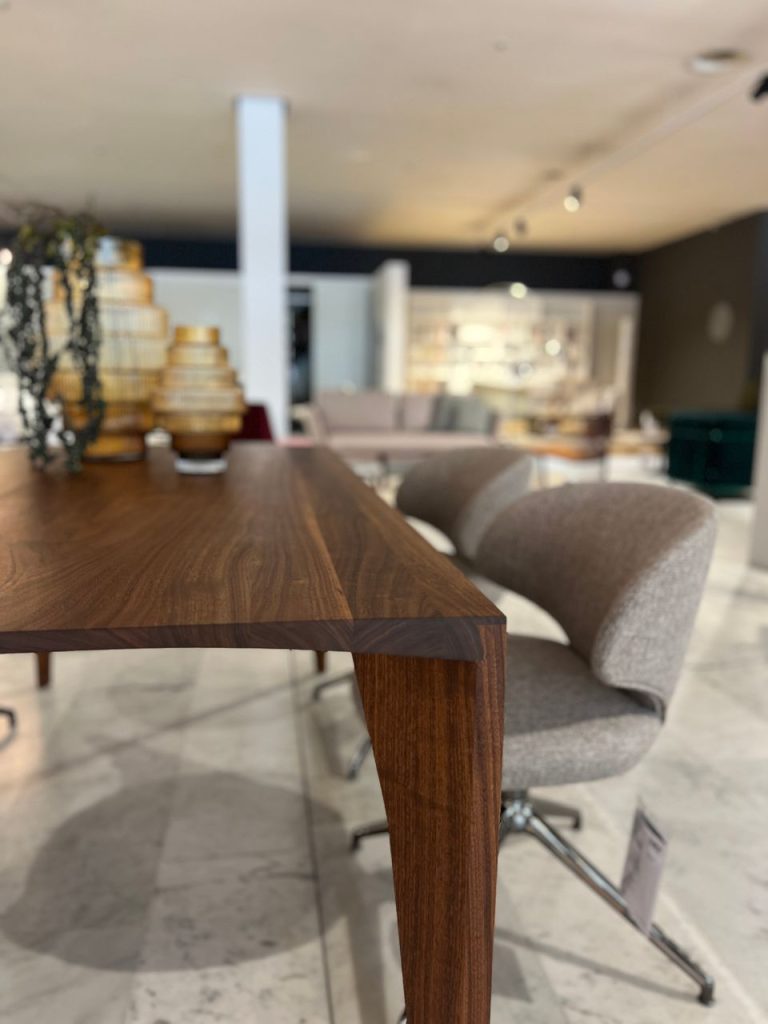 Close-up of a solid wood table edge in a modern showroom, with blurred chairs and display shelves in the background.