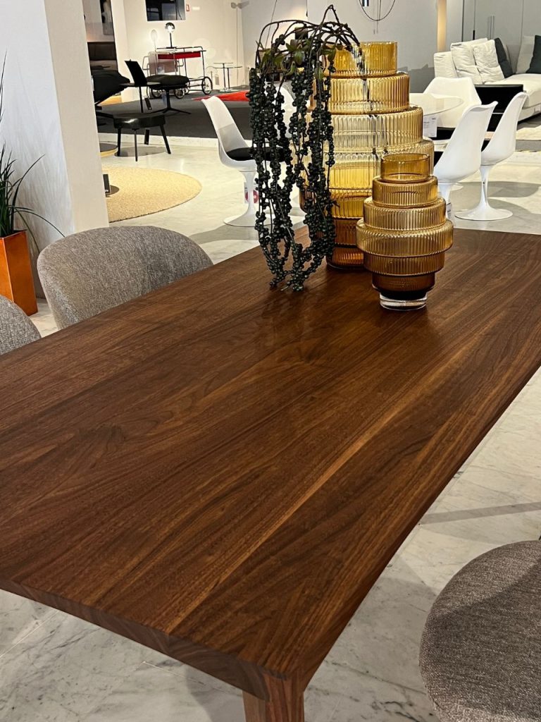 Wood dining table with stacked amber glass vases and a black bead garland centerpiece, set in a modern showroom with white chairs in the background.