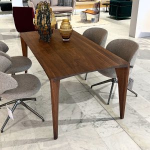 Rectangular walnut dining table with four tapered legs in a furniture showroom, surrounded by grey upholstered chairs.