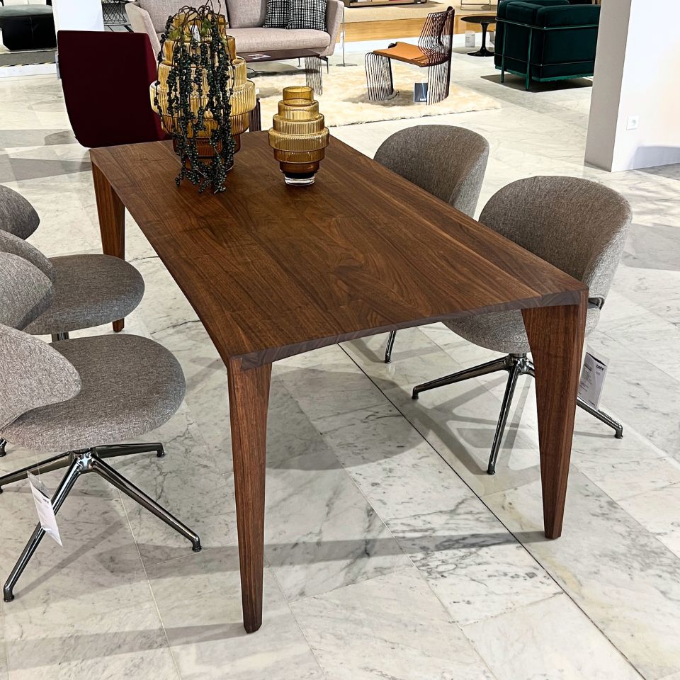 Rectangular walnut dining table with four tapered legs in a furniture showroom, surrounded by grey upholstered chairs.