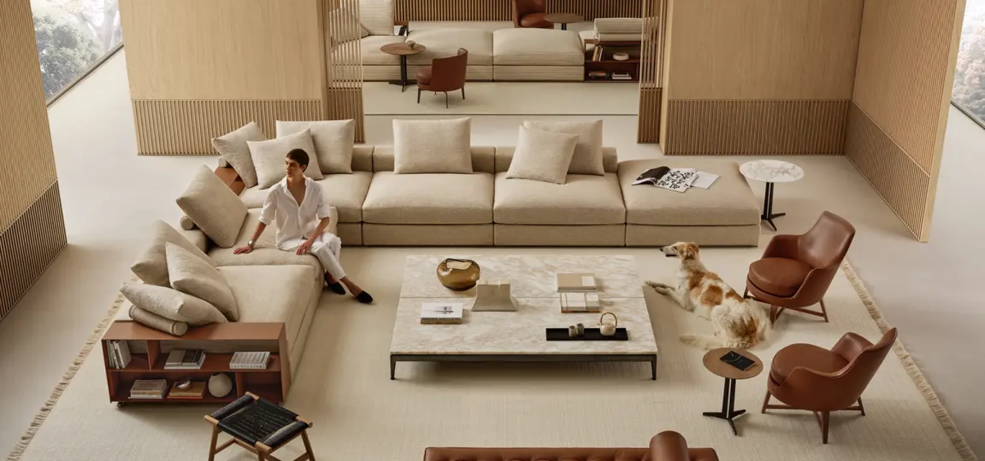 A modern beige living room with a large sectional sofa, a marble coffee table, and a dog near two brown leather chairs while a person sits on the sofa.