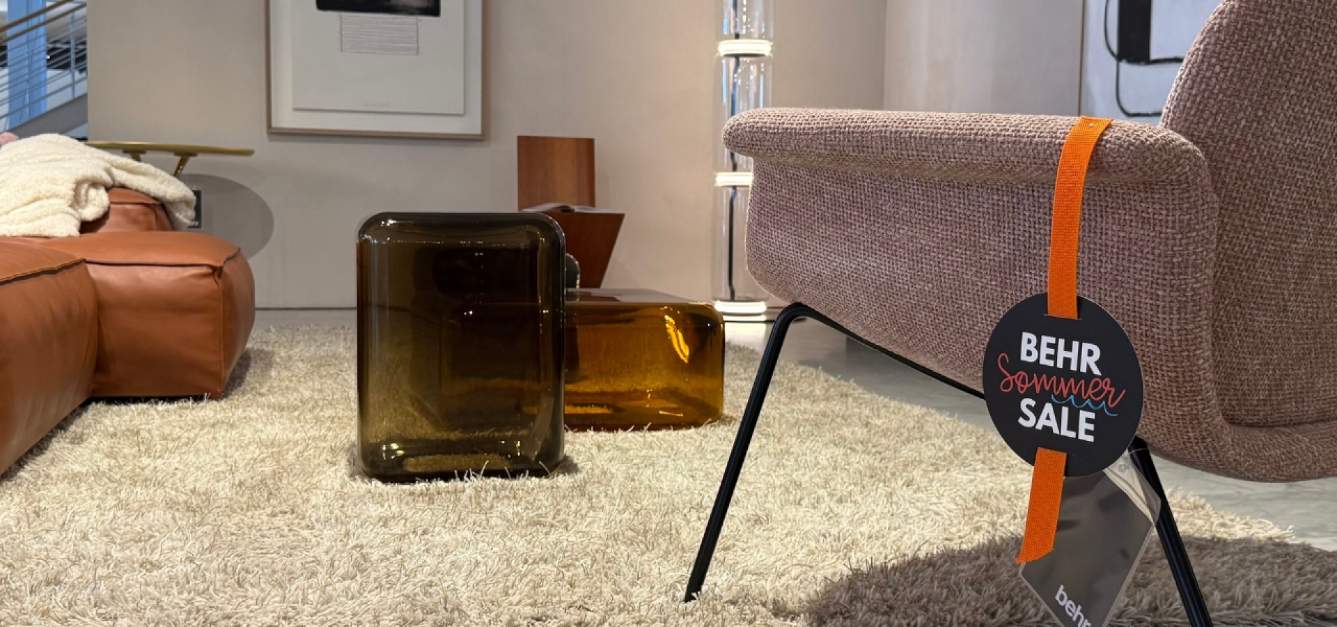 Showroom living room with a brown leather sectional, cream rug, and a beige fabric chair with an orange BEHR Summer Sale tag hanging from the arm.