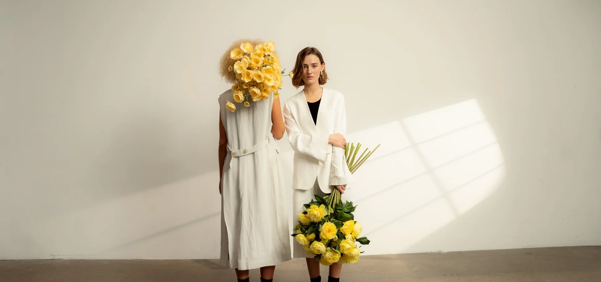 Two women in white outfits stand against a pale wall; the left person has a large yellow flower bouquet obscuring their head, while the right holds a cluster of yellow flowers and looks at the camera.